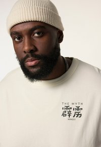 Krémové tričko s krátkým rukávem má černý grafický potisk s textem a symboly. Vzhled doplňuje žebrovaná, smetanově bílá čepice.