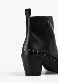 Botín negro de cuero hasta el tobillo con tacón cuadrado, que presenta tachuelas plateadas circulares a lo largo del costado y una lengüeta en la parte posterior para facilitar el uso.