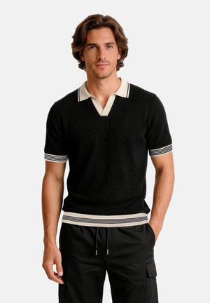 Uomo con capelli castani che indossa un maglione nero a maniche corte con colletto e polsini a righe bianche, abbinato a pantaloni neri con coulisse.