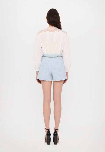 Camisa blanca de manga larga con detalles de encaje en la parte posterior, combinada con pantalones cortos de tweed azul claro con un dobladillo deshilachado y un cinturón de cadena plateada.