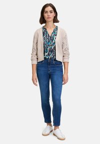 Beige gebreide cardigan, multicolored patroon blouse, blauwe denim jeans en witte sneakers met tan leren zolen; ontspannen pasvorm, lichte textuur in de stof.