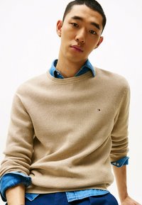 Beżowy sweter dzianinowy z teksturowanym wzorem, okrągłym dekoltem i detalem logo. Noszony na niebieskiej koszuli z dżinsu z zapiętymi mankietami.