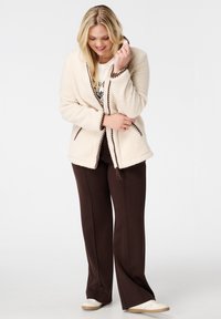 Crème-kleurige fleece jas met bruine afwerking, met een relaxte pasvorm en een open voorkant. Gecombineerd met bruine broek en witte schoenen.