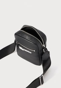 Borsa a tracolla in pelle nera con zip in argento, tasca frontale e tracolla regolabile. Presenta un dettaglio del logo sulla parte anteriore. Design compatto a forma quadrata.