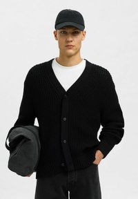 Jaunas vyras, vilkintis juodą megztinį su apykakle ant balto marškino, dėvi juodą kepurę ir laiko suvyniotą juodą maišą vienoje rankoje.