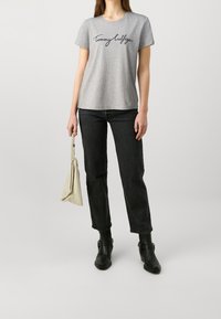 Vrouw in een grijs Tommy Hilfiger t-shirt, zwarte jeans en zwarte enkellaarsjes, met een beige handtas aan haar zijde.