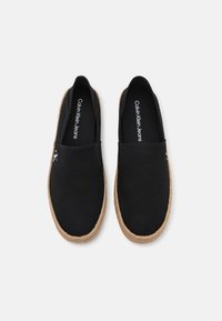 Chaussures noires à enfiler avec une tige en toile et une semelle en jute tissée. Présente un logo Calvin Klein Jeans à l'intérieur. Design minimaliste, sans motifs visibles.