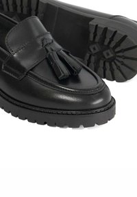 Black leather loafers featuring a front tassel, smooth surface, and rugged rubber sole with pronounced tread pattern for enhanced grip.
