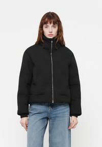 Svart polstret bomberjakke med høy krage og glidelås. Har ribbede mansjetter og en kort design. Båret med lyseblå vide jeans.