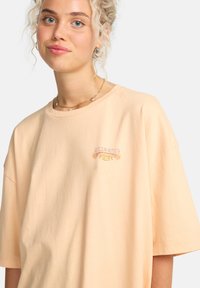 Φαρδιά κοντομάνικη μπλούζα από ελαφρύ ροδακινί ύφασμα με σχέδιο λογότυπου με λουλούδια σε παστέλ αποχρώσεις. Απαλή υφή από βαμβάκι.