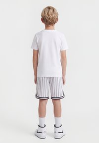 Dziecko z krótkimi blond włosami stoi tyłem, ubrane w prostą białą koszulkę, białe spodenki w cienkie paski, białe skarpetki i białe sportowe buty.