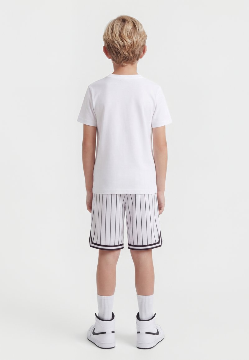 Dziecko z krótkimi blond włosami stoi tyłem, ubrane w prostą białą koszulkę, białe spodenki w cienkie paski, białe skarpetki i białe sportowe buty.