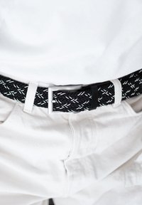 Gevlochten zwarte riem met witte stiksels, zilveren metalen gesp en zwarte leren accent, gedragen over witte broek.