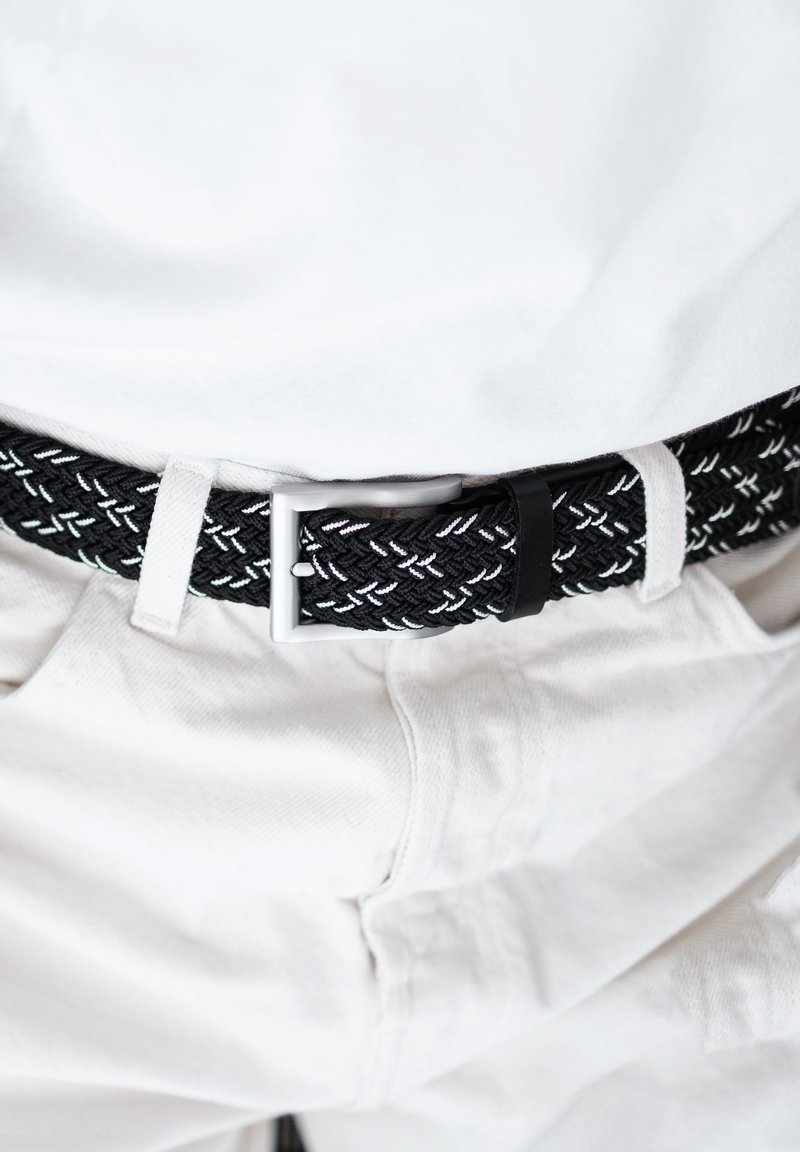 Gevlochten zwarte riem met witte stiksels, zilveren metalen gesp en zwarte leren accent, gedragen over witte broek.