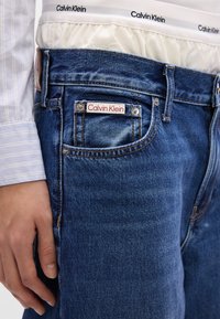 Ciemnoniebieskie dżinsy z dodatkiem czerwono-białej naszywki z logo "Calvin Klein", widocznymi metalowymi elementami i przetartą fakturą.