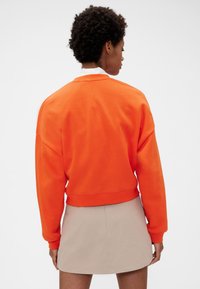 Oranžová zkrácená mikina s kulatým výstřihem a spadenými rameny, kombinovaná s béžovou sukní. Hladká bavlněná textura.