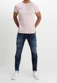Camiseta de algodón de color rosa claro con cuello redondo, combinada con jeans de mezclilla azul oscuro y zapatillas blancas. Diseño sencillo, mangas cortas y forma ajustada.