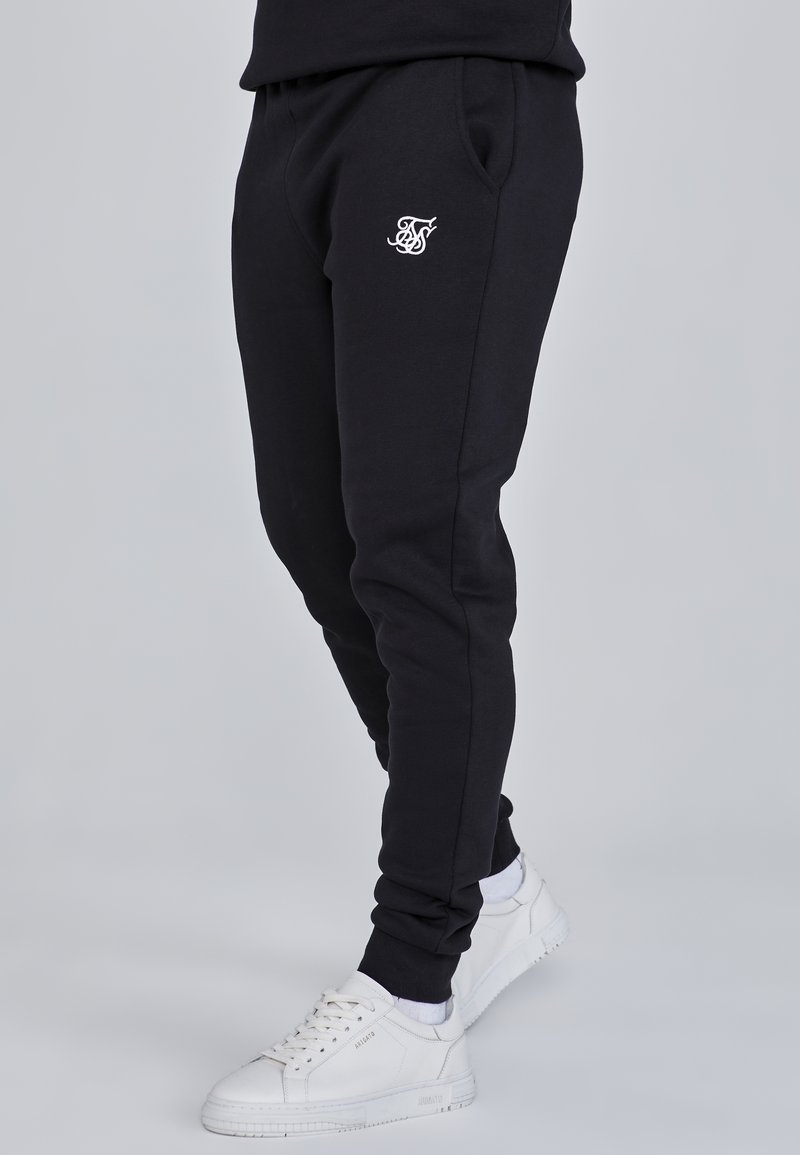 Zwarte joggingbroek van zacht materiaal, met zijzakken en een geborduurd logo in het wit op de linker dij. Gemaakt in combinatie met witte sneakers.
