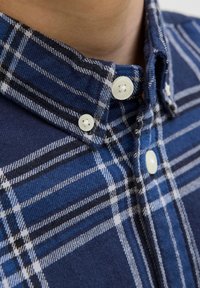 Detailný záber na golier modrej károvanej košele s bielymi gombíkmi, zobrazujúci detailnú textúru látky a stehy na gombíkoch.