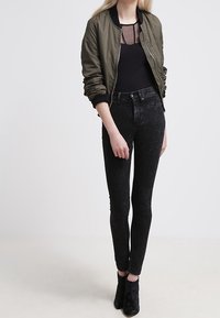 Nainen, joka on pukeutunut oliivinvihreään bomber-takkiin, mustaan verkkopaitaan, korkeavyötäröisiin haalistuneisiin mustiin skinny-farkkuihin ja mustiin nilkkureihin, seisoo valkoista taustaa vasten.