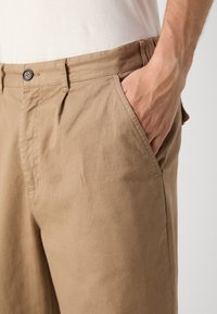Khakifarbene Baumwollhose mit einer lockeren Passform, seitlichen Taschen und einem Knopfverschluss vorne, mit einer glatten Textur und minimalen Falten.