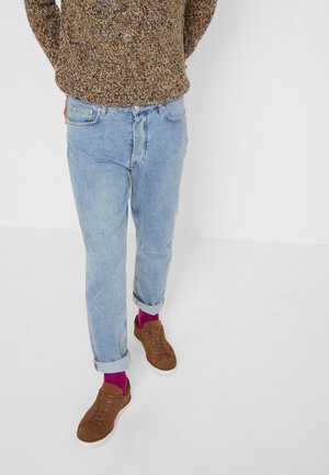 Person mit hochgekrempelten hellblauen Jeans, braunen Sneakers, knallpinken Socken und einem mehrfarbigen Strickpullover steht vor einem weißen Hintergrund.