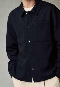 Černá prošívaná bunda se dvěma kapsami na hrudi, předními zapínáními na druky a límcem. Látka má strukturovaný povrch.