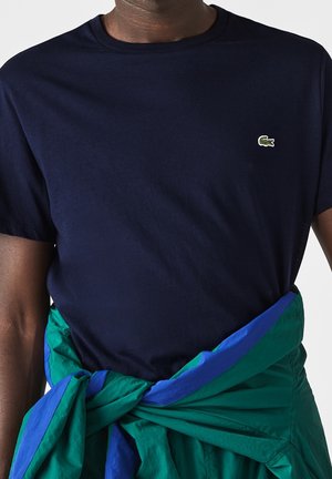 Granatowy bawełniany t-shirt z okrągłym dekoltem i małym zielonym haftowanym logo. Zielono-niebieska kurtka związana jest w talii.