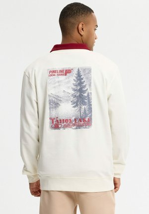Joven con sudadera blanca con cuello rojo y escena impresa de la naturaleza del lago Tahoe en la espalda, de pie de espaldas.