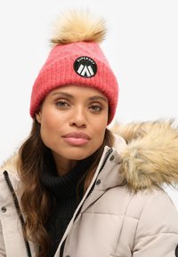 Gorro de punto rosa con un pompón de piel sintética y un logo redondo negro de "Superdry". Usado con un abrigo beige que tiene una capucha con borde de piel.
