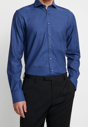 Man draagt een nauwsluitend blauw overhemd met lange mouwen en knopen, ingestopt in een zwarte broek, tegen een effen achtergrond.