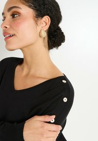 Mujer con cabello rizado oscuro recogido en un moño, lleva un suéter negro de cuello en V con tres botones en el hombro y pendientes en forma de lágrima beige.