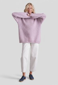 Blondynka w za dużym lawendowym swetrze, białe spodnie o kroju capri i granatowe trampki na jednolitym szarym tle.