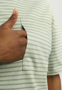 Pruhované svetlozelené tričko s otvorenou kapsou na ľavej strane; ruka je čiastočne viditeľná vo vnútri vrecka. Mäkkučký textil z bavlny.