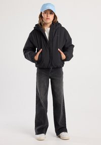 Black puffer jacket with hood, zip closure, and elastic cuffs. Worn over a light shirt and paired with faded black wide-leg jeans and white sneakers. Light blue cap.