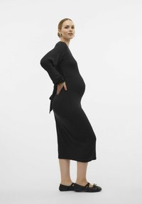 Vestito premaman nero a coste con maniche lunghe, dettaglio di laccio laterale, orlo fino alla caviglia e scarpe nere basse. Design semplice, aderente.