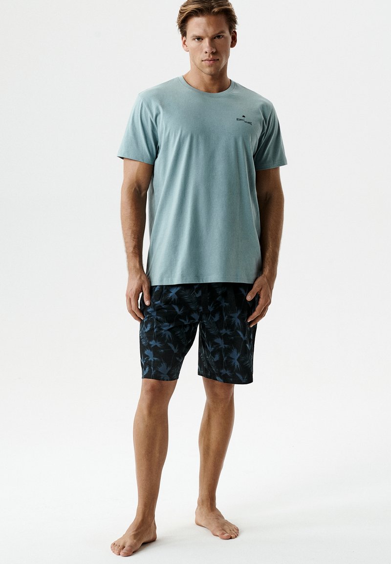 Młody mężczyzna stojący boso, ubrany w jasnoturkusową koszulkę i czarne szorty z niebieskimi kwiatowymi wzorami na białym tle.