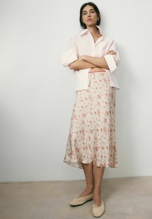 Frau steht mit verschränkten Armen, trägt ein hellrosa Shirt, einen floralen Midirock und beige Ballerinas vor einem schlichten Wandhintergrund.