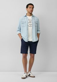 Lichtblauw denim overhemd over een wit graphic t-shirt, marineblauwe shorts en beige sneakers met bruine accenten; casual, getailleerde pasvorm.