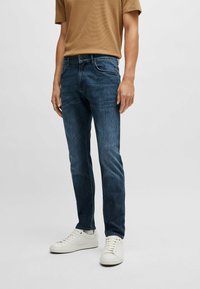 Jeans en denim bleu avec une coupe slim, présentant un design classique à cinq poches, un léger délavage et une fermeture à glissière. Associés à des baskets blanches.