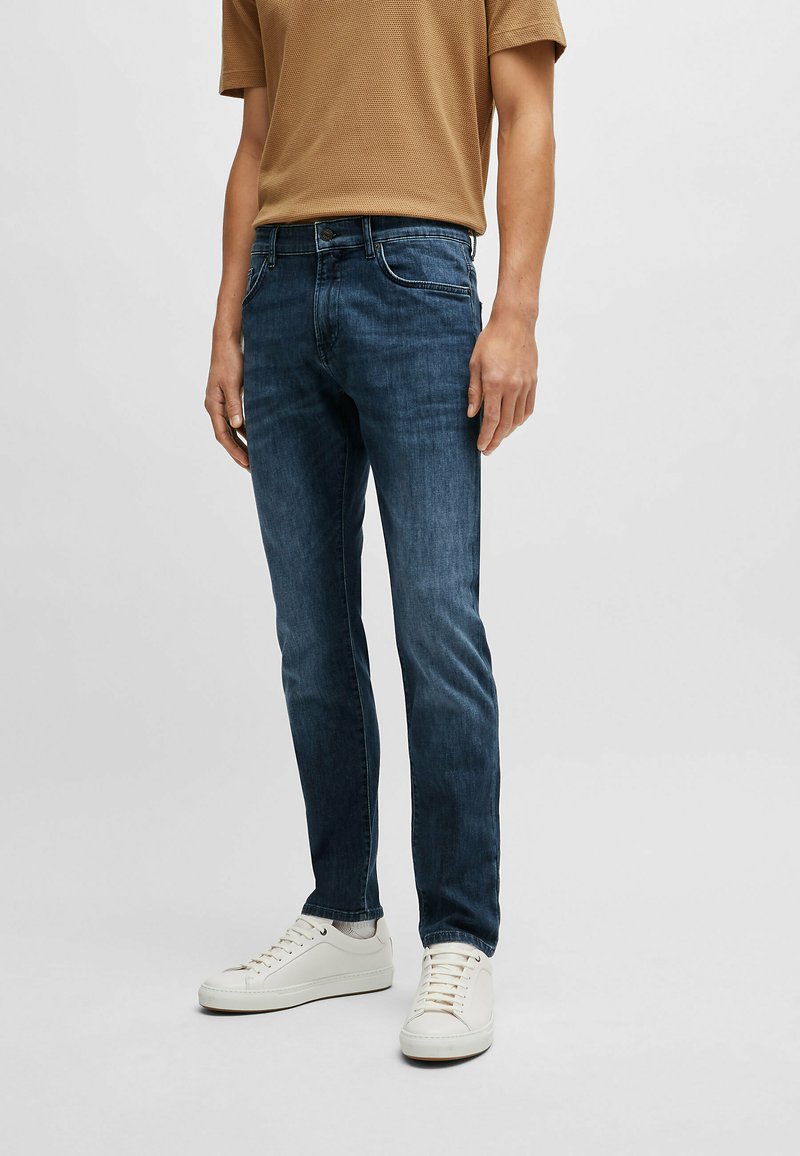 Jeans en denim bleu avec une coupe slim, présentant un design classique à cinq poches, un léger délavage et une fermeture à glissière. Associés à des baskets blanches.