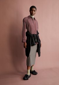 Long-sleeve, burgundy striped shirt, tied black jacket at waist, grey pencil skirt, green socks, and black detailed footwear. Simple background.