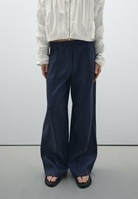 Personne portant un pantalon large bleu foncé, une veste blanche zippée et des sandales noires, se tenant sur un sol gris clair devant un mur gris uni.