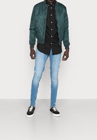 Tmavě zelená bomber bunda, černá košile s knoflíky, světle modré slim-fit džíny a černé tenisky s bílou podrážkou. Hladké a lehké materiály.