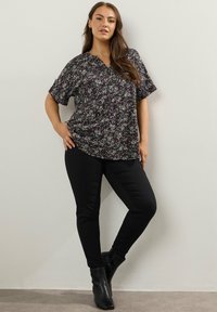 Donna in piedi contro un muro che indossa una blusa corta a maniche corte con fantasia floreale scura, pantaloni neri aderenti e stivali neri alla caviglia.
