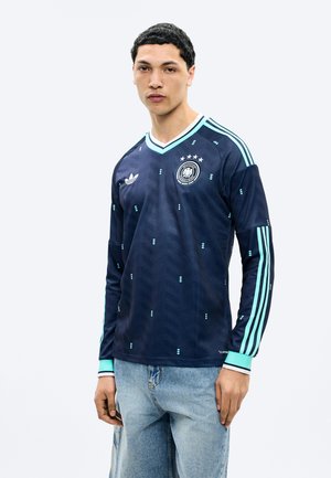 Νεαρός άνδρας που στέκεται φορώντας μπλε σκούρα αθλητική μπλούζα με μακριά μανίκια, με ανοιχτό μπλε ρίγες και έμβλημα στο στήθος, συνδυασμένη με ανοιχτό μπλε τζιν παντελόνι.