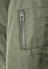 Grön tygjacka med detalj av en zippered ficka med metallblixtlås, strukturerad tyg och synlig sömnad vid kanterna.