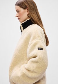 Μπεζ φλις ζακέτα με ψηλό μαύρο γιακά, με έντονο λογότυπο πάνω στο μανίκι και μαλακή, υφασμάτινη επιφάνεια.