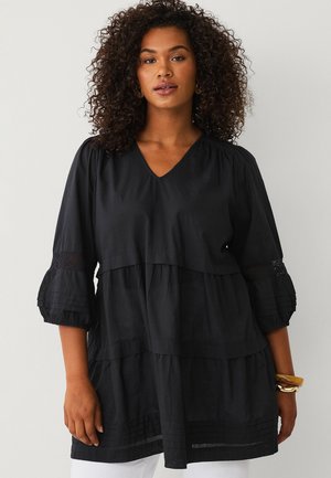 Vrouw met krullend haar die een losse zwarte V-hals blouse draagt met gelaagde lagen en driekwart mouwen en een houten armband om haar pols.