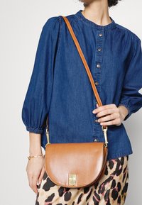 Donna che indossa una camicetta blu con bottoni e una gonna a stampa leopardo, mentre tiene in mano una borsa a tracolla in pelle color beige con dettagli in oro sulla chiusura e sulla tracolla.